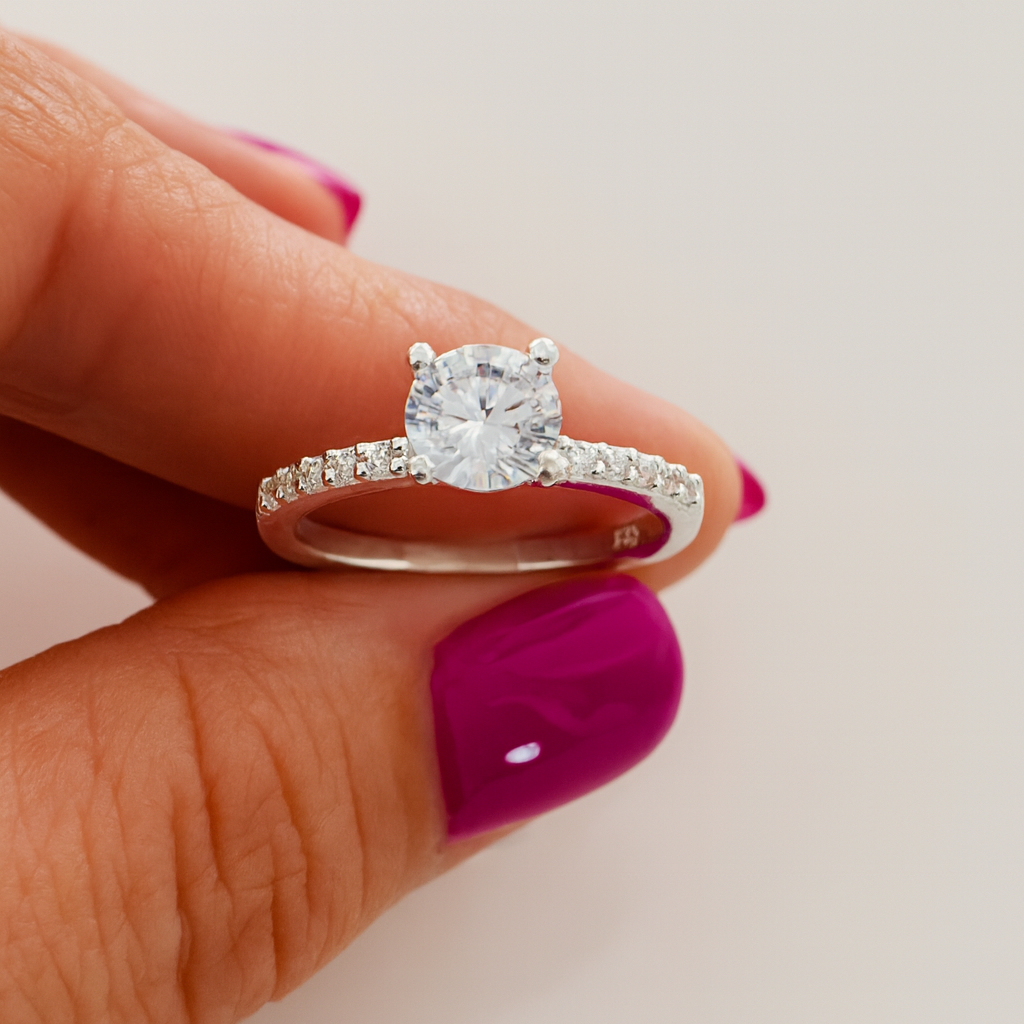 nel de logodnă din argint cu un diamant rotund central și pietre mici pe bandă, ținut între degete cu manichiură violet, pe fundal alb curat.