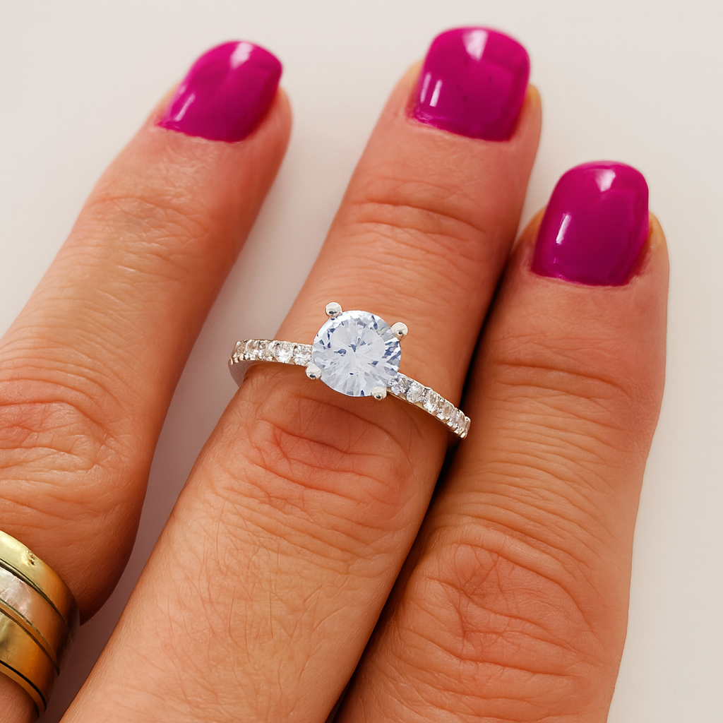 nel de logodnă elegant din aur alb, cu diamant rotund central și pietre mici pe bandă, purtat pe degetul inelar al unei mâini cu manichiură fucsia.